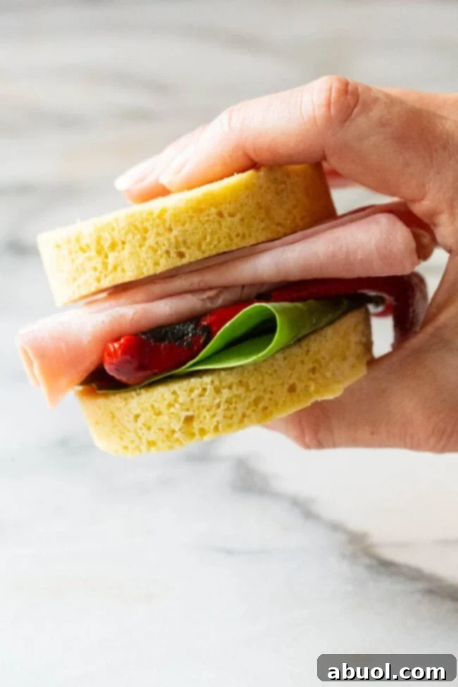 A hand holds a small, enticing sandwich made with two thick slices of low-carb bread. The filling includes savory deli meat, vibrant roasted red pepper, and fresh spinach leaves, showcasing the bread's versatility.