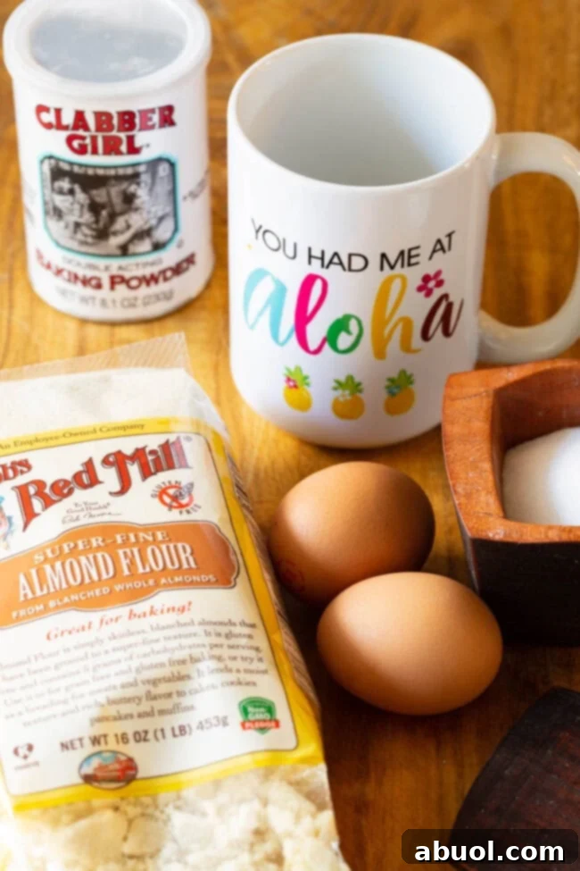 A beautifully arranged flat lay showing the simple ingredients for low-carb bread: a white mug, a container of grain-free baking powder, a bag of fine-ground almond flour, two fresh eggs, and a small wooden bowl of sea salt.