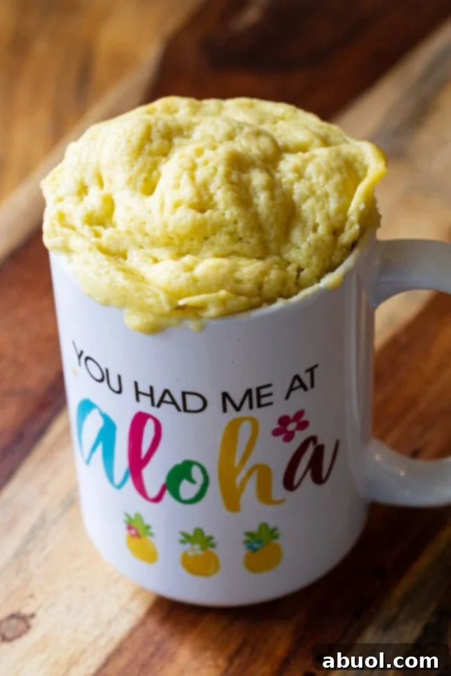A perfectly puffed, golden-brown loaf of microwave bread is gently popping out from the top of a white mug, showcasing its light and airy texture.
