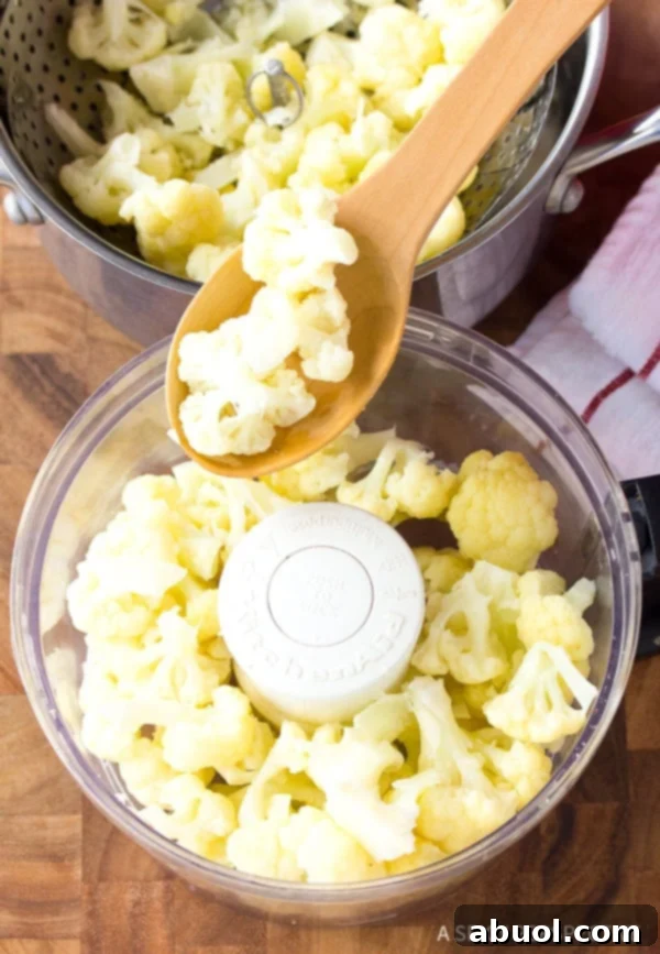 Wood spoon adding steamed cauliflower florets to a food processor.