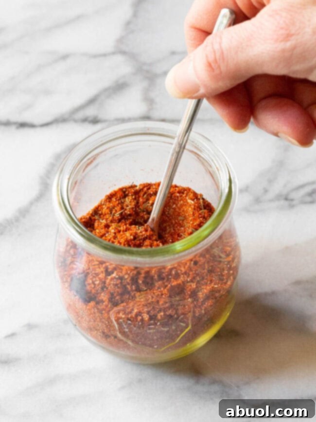 A hand holding a spoon dipped into a glass jar filled with vibrant homemade blackened seasoning, ready for use.