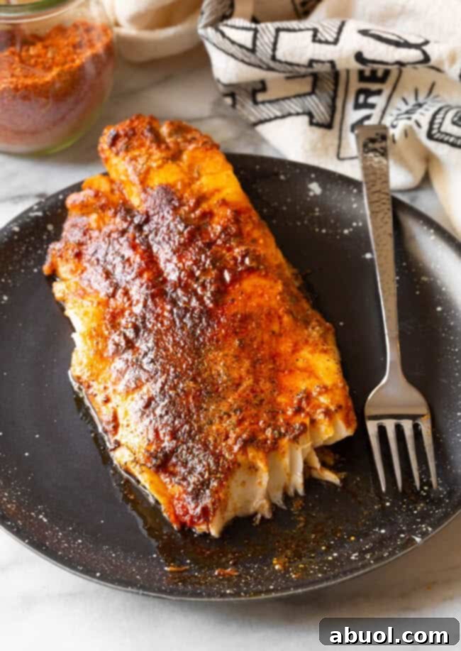 A perfectly cooked fillet of blackened fish, coated in a dark, flavorful crust, served on a sleek black plate alongside a metal fork.