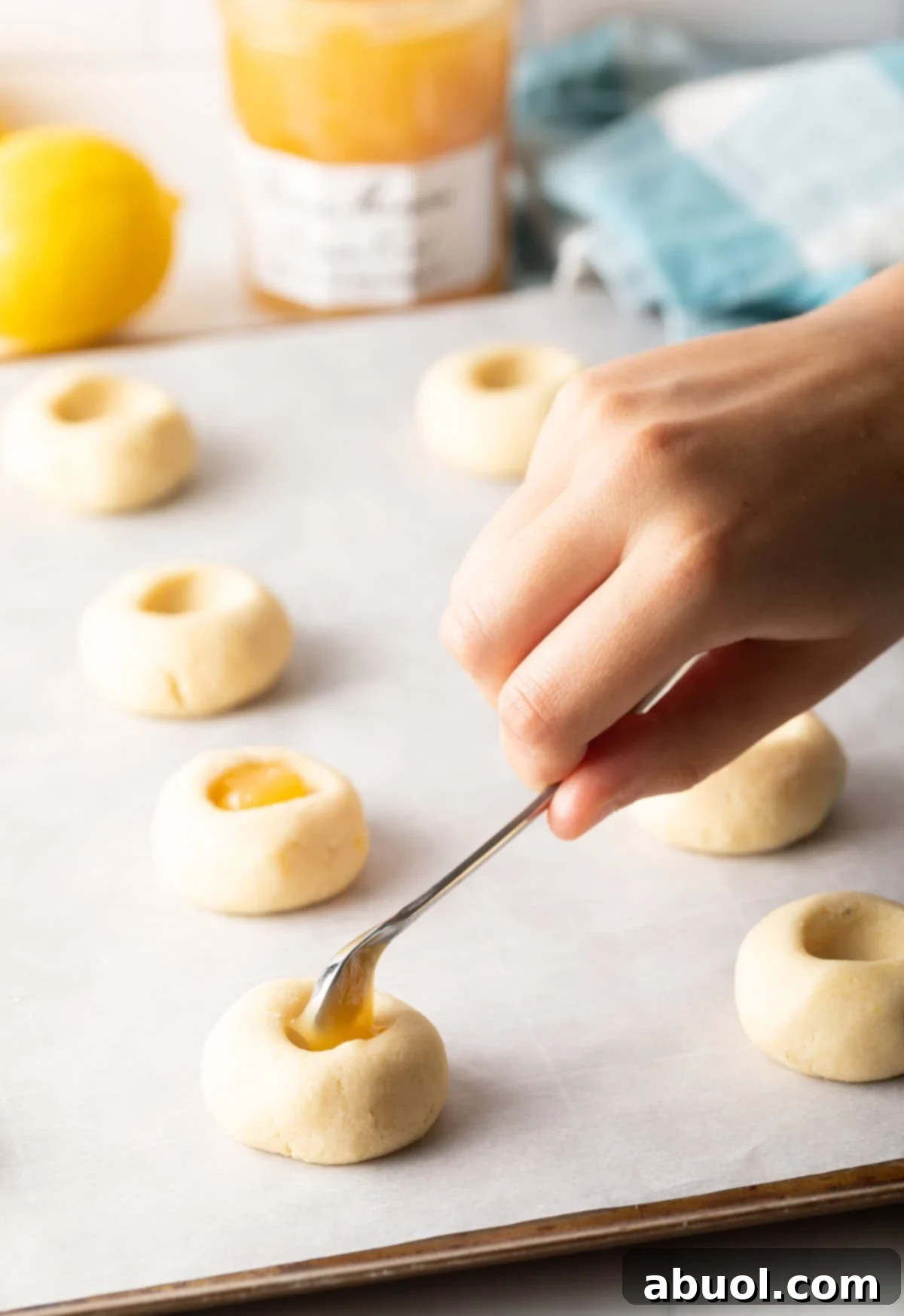 Close-up of a small spoon carefully dolloping lemon custard into the indent of a thumbprint cookie dough.