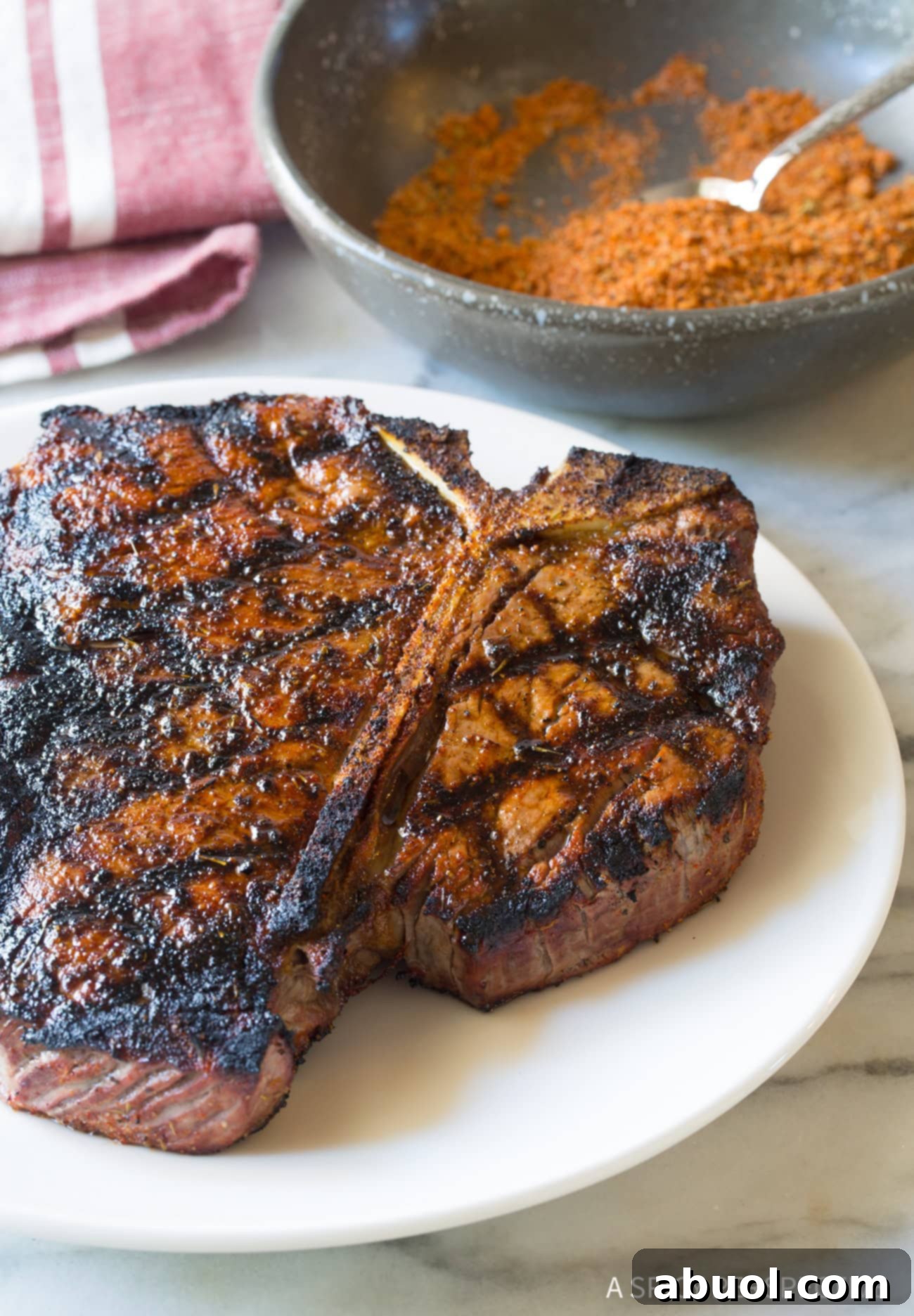 A succulent grilled steak on a white plate with a small bowl of steak seasoning in the background, showcasing its delicious appearance.