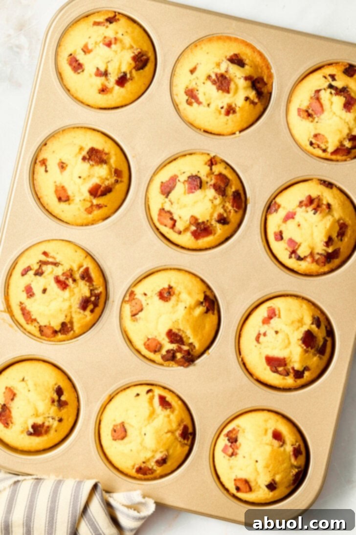 Heavenly Bacon Corn Muffins 10 Freshly baked corn muffins with bacon bits visible, cooling in a muffin pan, drizzled with maple syrup.