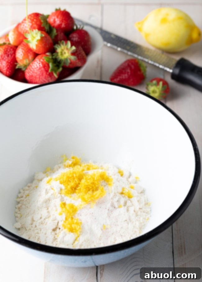 Flour and lemon zest in a mixing bowl with a bowl of strawberries beside it.