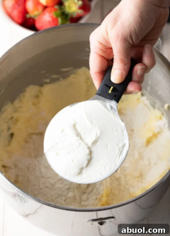 Strawberry cake batter in a mixing bowl with a hand holding a measuring cup with yogurt above it.