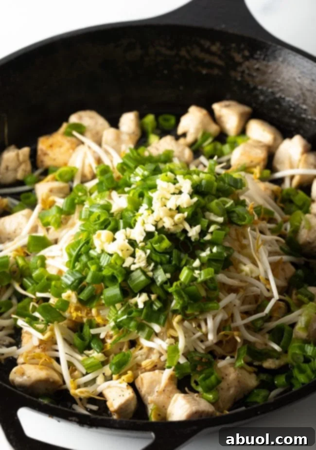 A dynamic shot showing fresh mung bean sprouts, vibrant chopped green onions, and aromatic minced garlic being added to the large skillet with perfectly cooked chicken pieces, beginning the aromatic stir-fry.