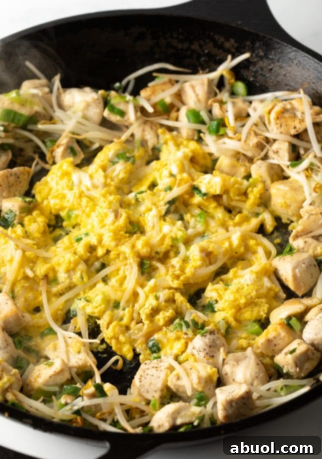 A close-up view of fluffy scrambled eggs being incorporated into the sautéed mixture of chicken, mung bean sprouts, green onions, and garlic in a large skillet, adding richness to the Pad Thai base.