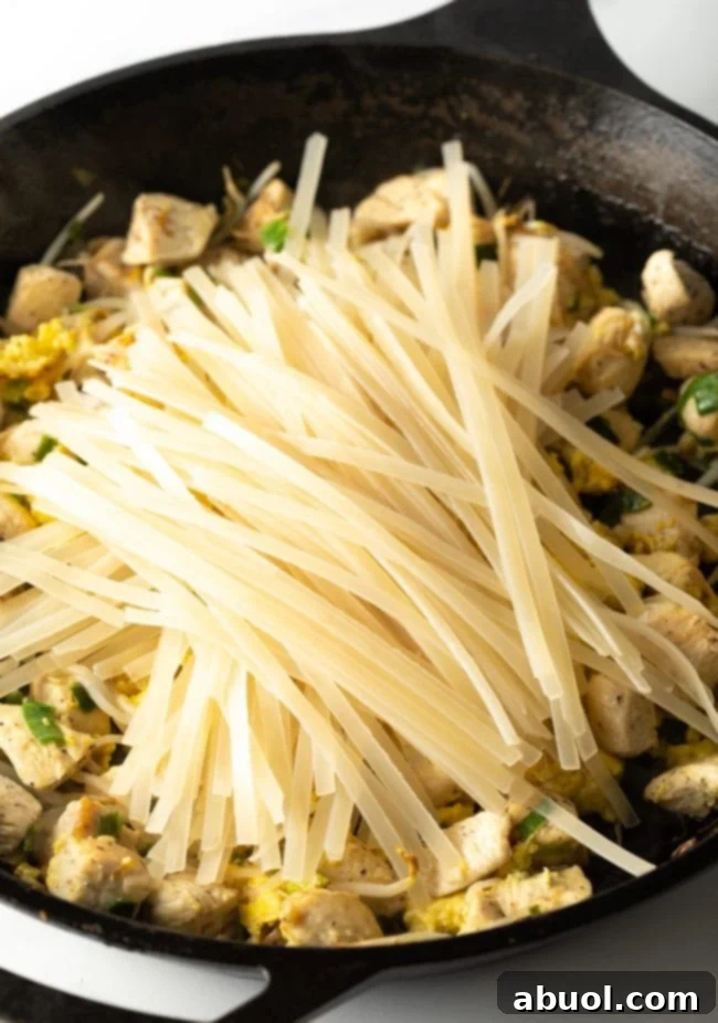 The moment perfectly soaked rice noodles are added on top of the cooked chicken, scrambled eggs, bean sprouts, green onions, and garlic in a large skillet, preparing for the final mix with the sauce.