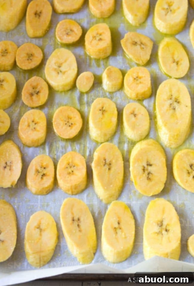 Raw, diagonally sliced plantains meticulously arranged on a parchment-lined baking sheet, awaiting their transformation in the oven.