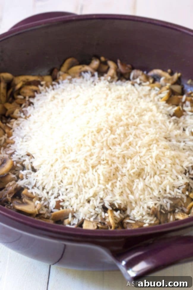 A pan with sautéed mushrooms and aromatics, topped with uncooked long-grain rice, poised to absorb the rich broth and transform into a luscious pilaf.
