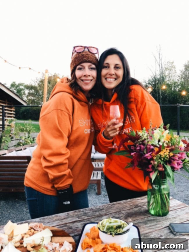 Two women in orange SkinnyTaste hoodies, one holding a wine glass, enjoying the glamping trip in Ithaca.