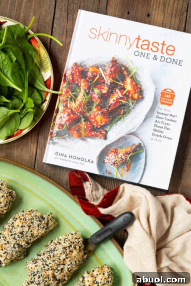 The SkinnyTaste One & Done Cookbook open on a table next to a plate of air fryer chicken roll-ups and a fresh bowl of spinach, showcasing a healthy meal.