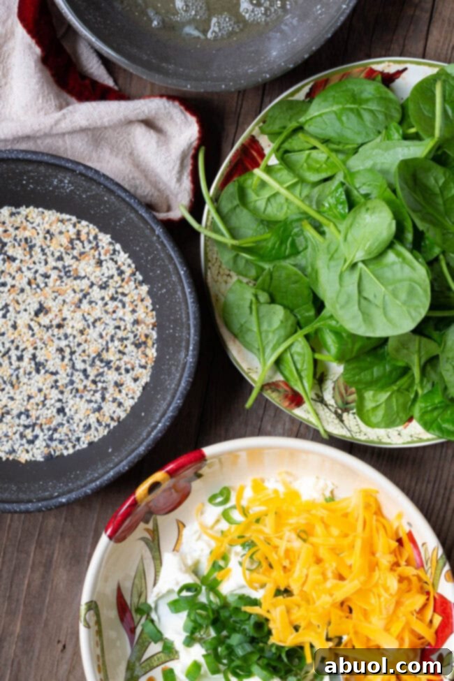 Close-up of recipe ingredients including a bowl of fresh baby spinach, a shallow dish with everything bagel seasoning, and a bowl containing a creamy mixture of softened cream cheese, chopped green onions, and shredded cheddar cheese.