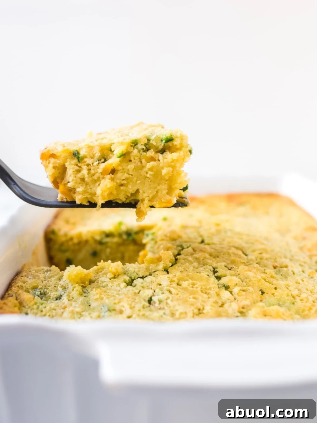 Side view of casserole dish and a spatula holding a piece of corn pudding, showcasing its golden-brown top and tender interior.