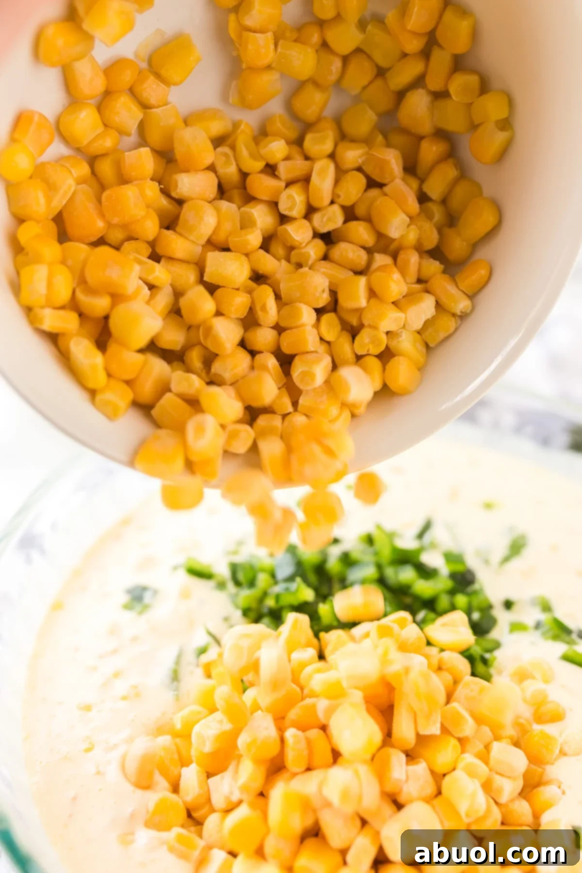 Pouring a bowl of fresh corn kernels into another bowl containing a creamy mixture of other ingredients, preparing the corn pudding batter.
