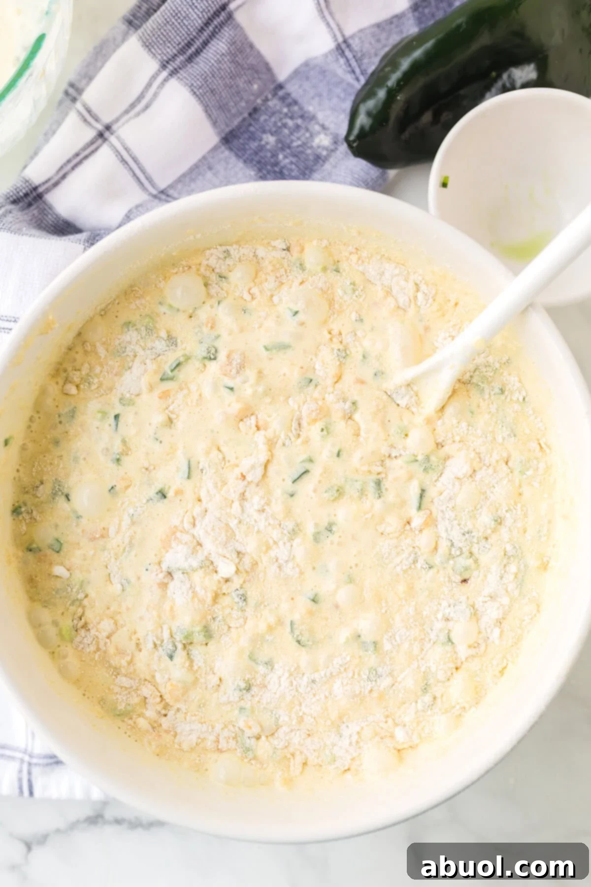 Top view of a bowl containing the perfectly mixed batter for the corn pudding, with a spoon resting in it, ready for the next step.