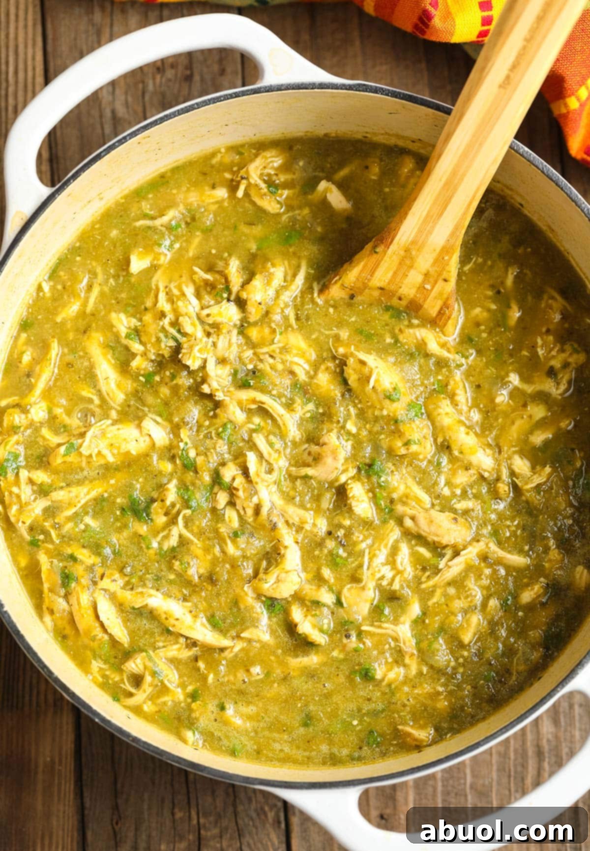 Top view of a large sauce pot filled with simmering Chicken Chili Verde, showcasing its rich green color and tender chicken shreds. A wooden spoon rests in the pot, ready to serve. 