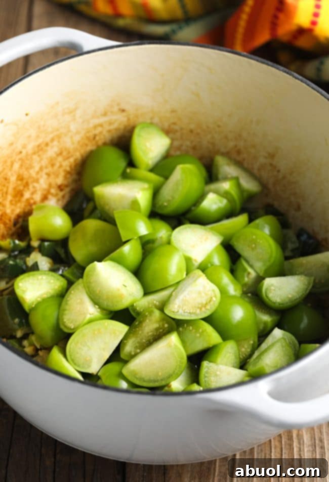 Quartered tomatillos added to a pot with sautéed onions, garlic, and peppers, ready to release their tangy essence.