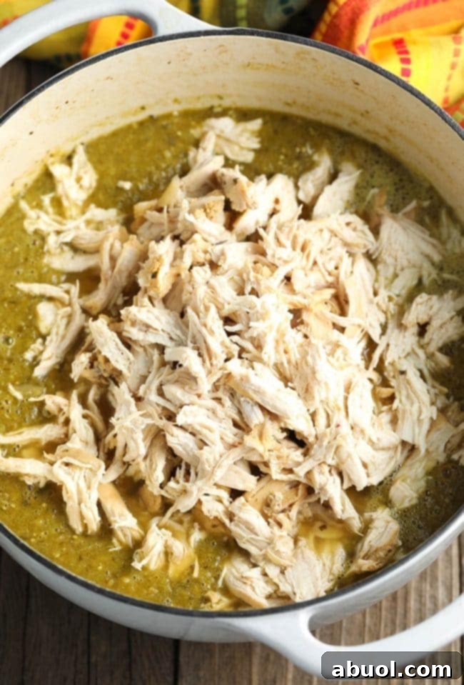 Cooked and shredded chicken pieces being gently stirred back into the pureed green chili base, ensuring every strand is coated.
