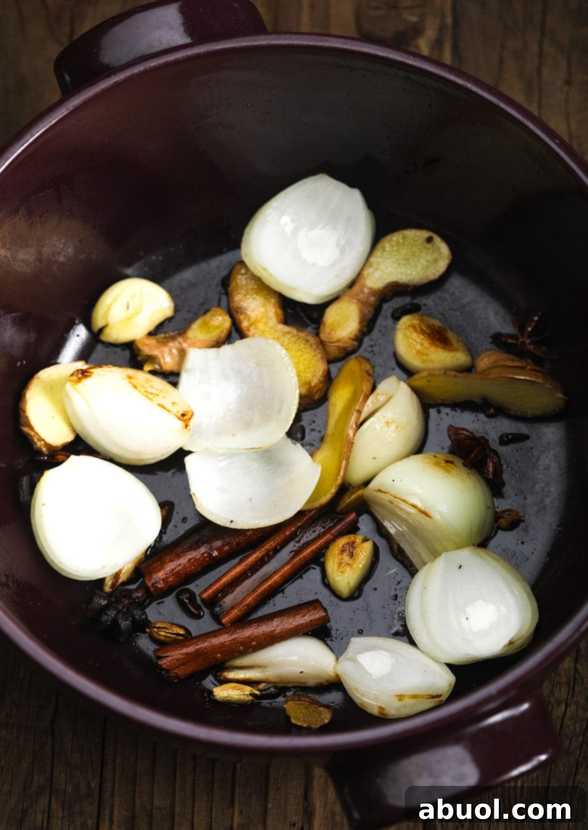 A large pot on a stovetop, showing the aromatic foundation of pho broth with quartered onions, slices of ginger, whole garlic cloves, and cinnamon sticks being sautéed.