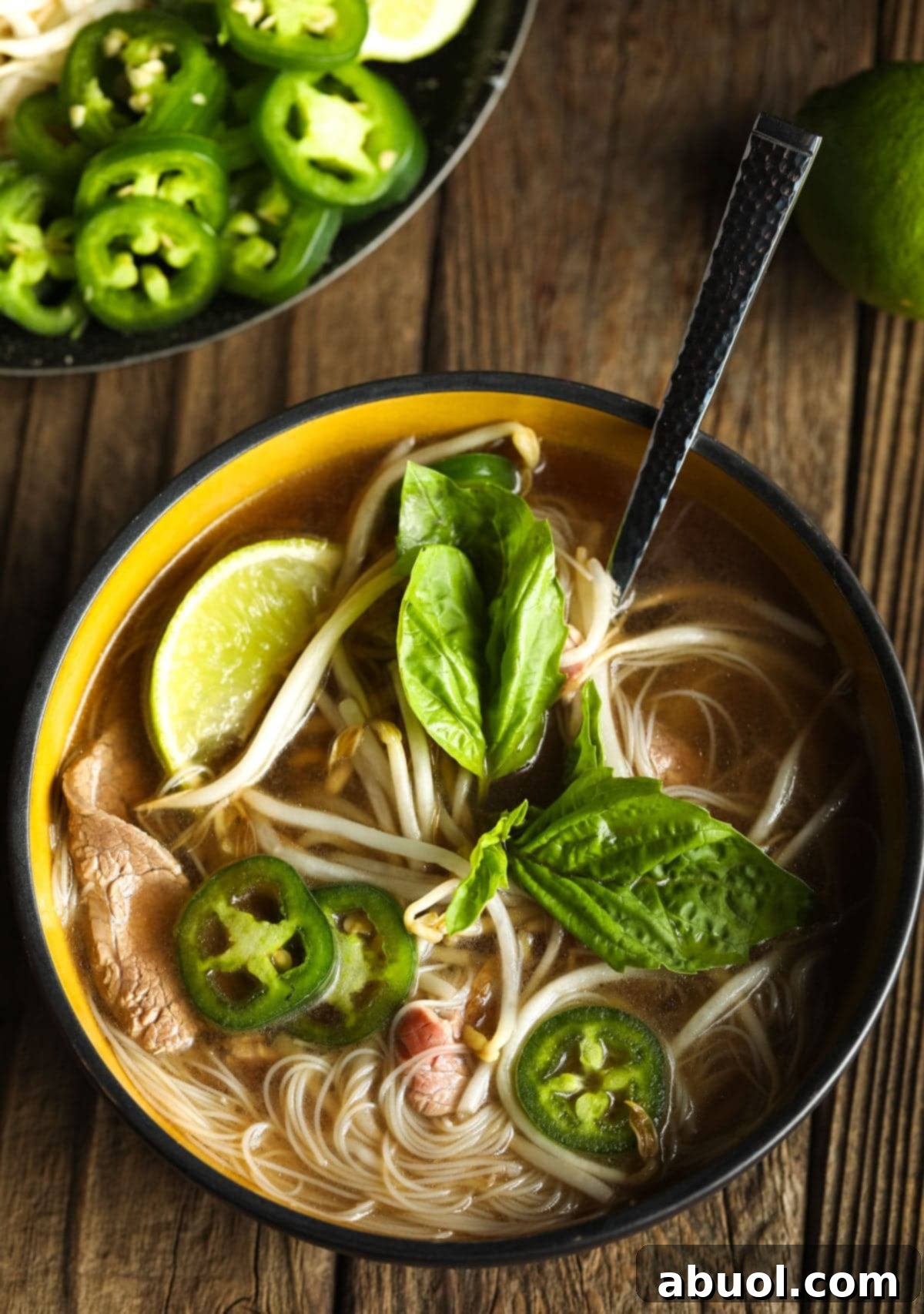 An overhead shot of a prepared bowl of pho, beautifully garnished with fresh basil leaves, sliced jalapeños, and lime wedges, adding color and flavor to the aromatic soup.