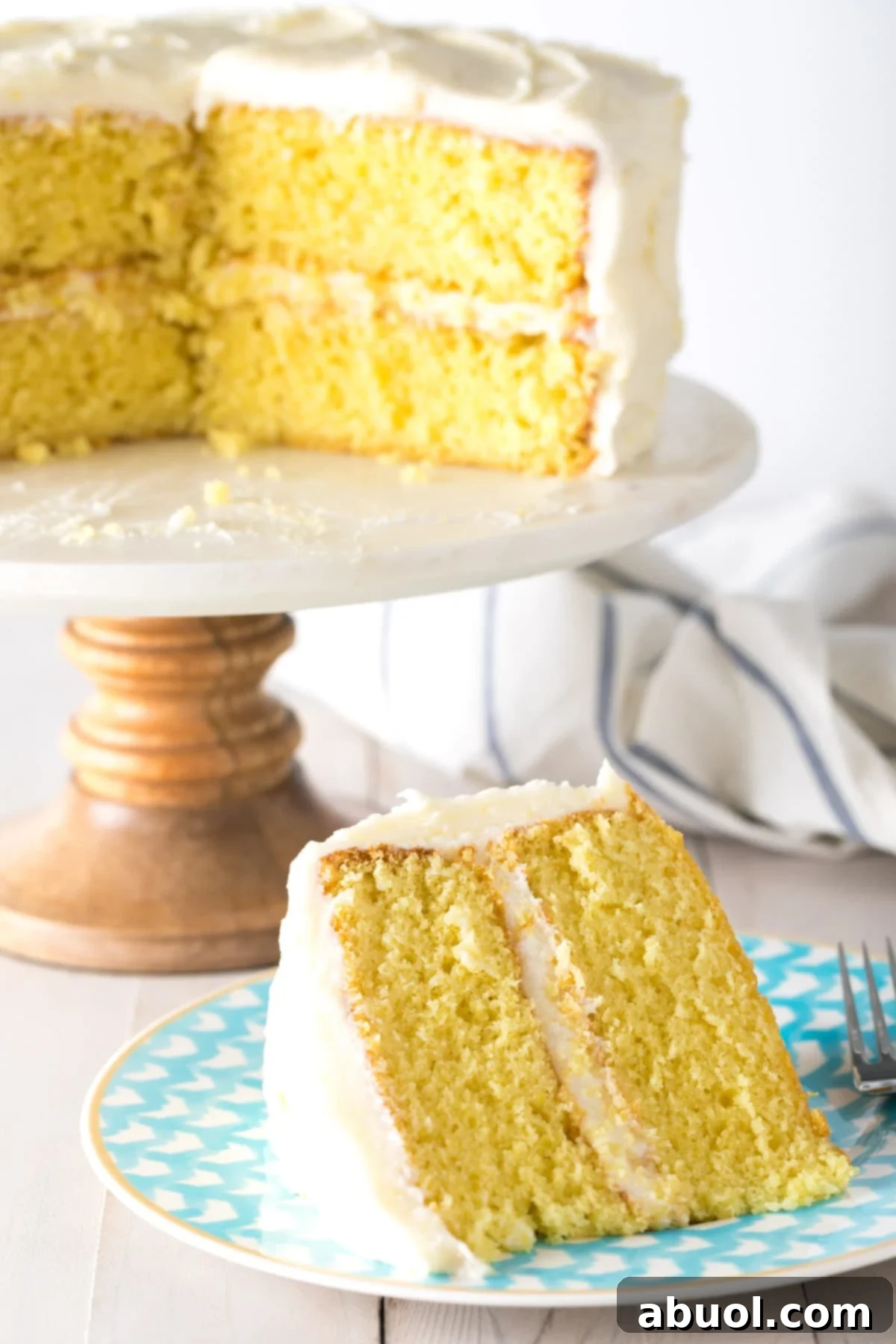 A slice of lemon buttermilk cake on a blue plate, with the rest of the cake on a stand in the background.