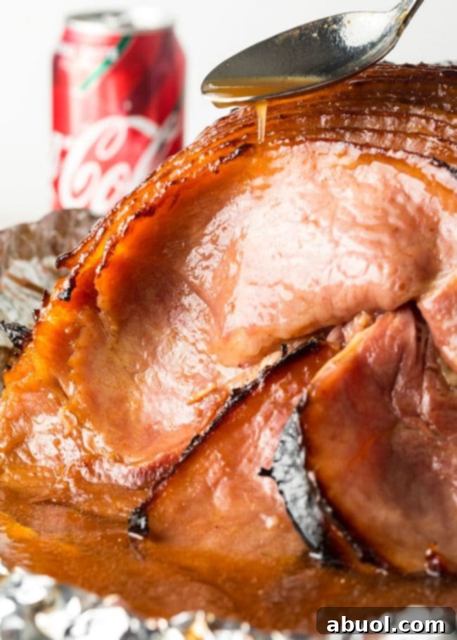 A hand using a spoon to spread the glistening Coca-Cola glaze over the top of the baked ham.
