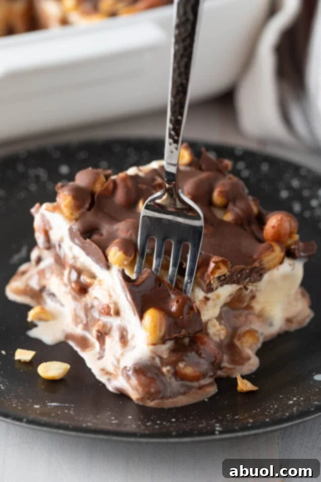 Close-up of a slice of Buster Bar Ice Cream Cake on a plate, with a fork poised to take a bite, showing the distinct layers of the recipe.