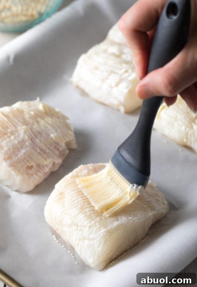 A hand using a brush to apply a thin layer of mayonnaise to a raw fish fillet, preparing it for seasoning.