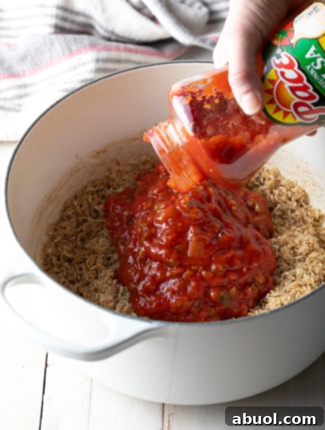 Chunky tomato salsa being poured into a pot with sautéed long-grain white rice, ready to be cooked into Spanish rice.