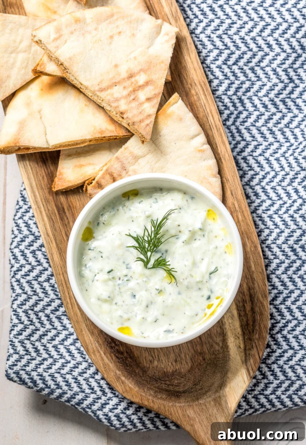 Creamy Tzatziki dip in a white bowl, elegantly placed on a rustic wood platter alongside warm pita bread triangles, ready for dipping.