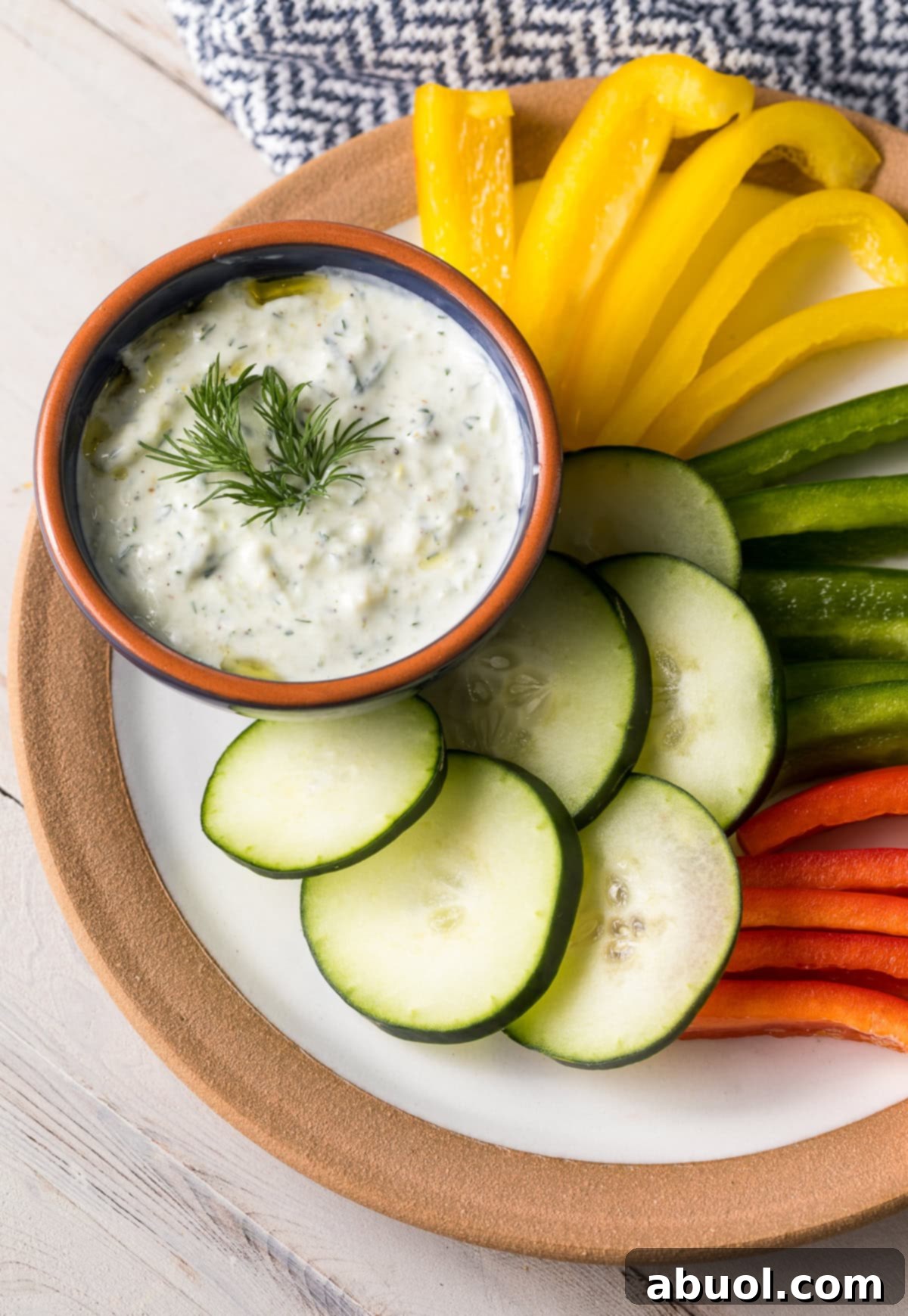 A vibrant display of tzatziki dip in an orange and blue patterned bowl, nestled on a rustic serving platter next to an assortment of colorful cut-up vegetables like bell peppers, cucumbers, and carrots, perfect for dipping.