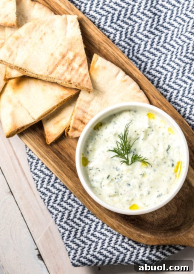 Freshly prepared Tzatziki dip in a white ceramic bowl, garnished with a sprig of dill, resting on a textured wooden board next to several triangles of warm pita bread.