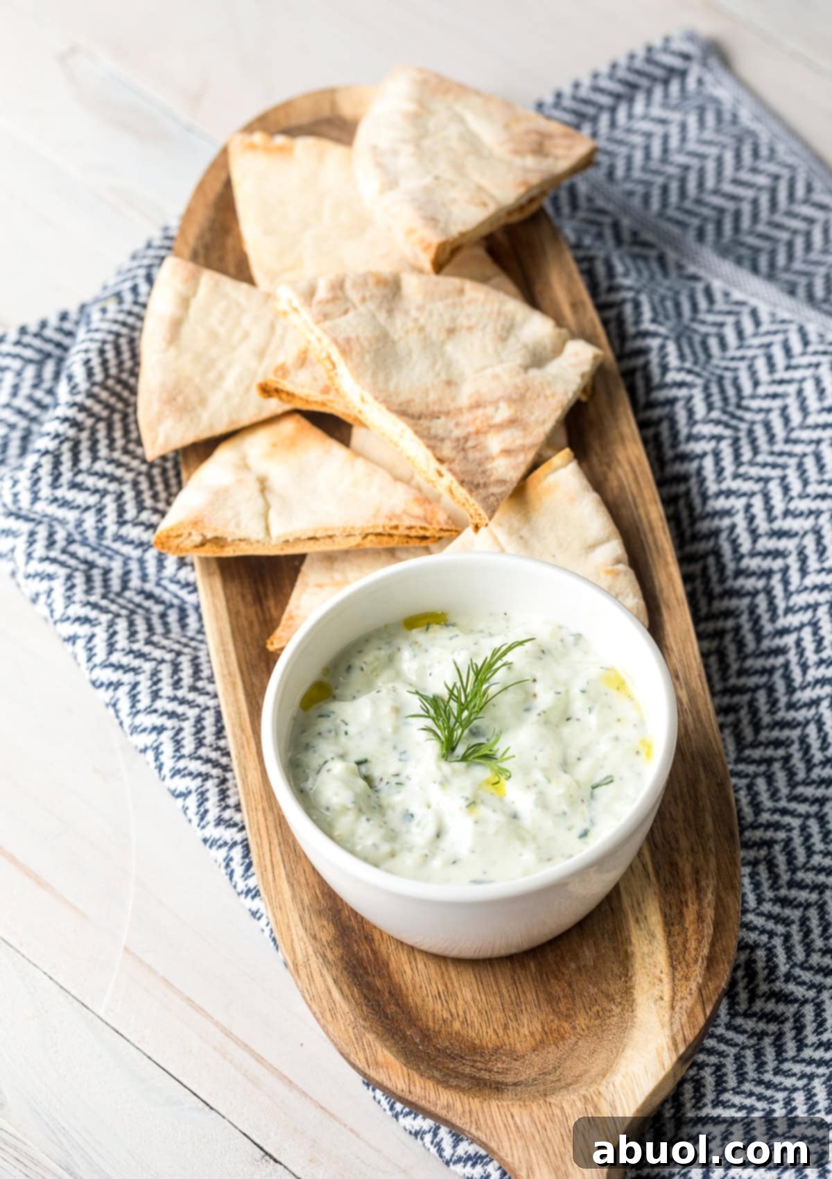 A close-up shot of creamy homemade Tzatziki dip in a white bowl, garnished with fresh dill, placed on a light wooden platter with a side of warm pita bread.