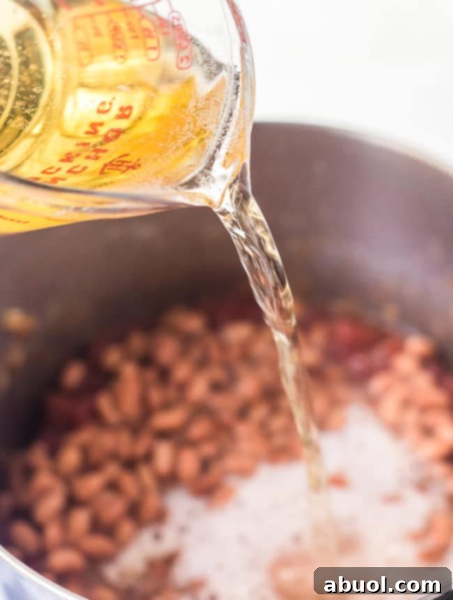 A measuring cup carefully pouring beer into a stock pot filled with aromatic chili ingredients.