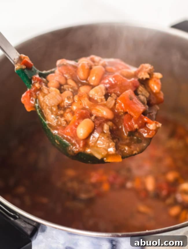 A ladle filled with rich steak chili with beer, with the full pot of simmering chili blurred in the background.