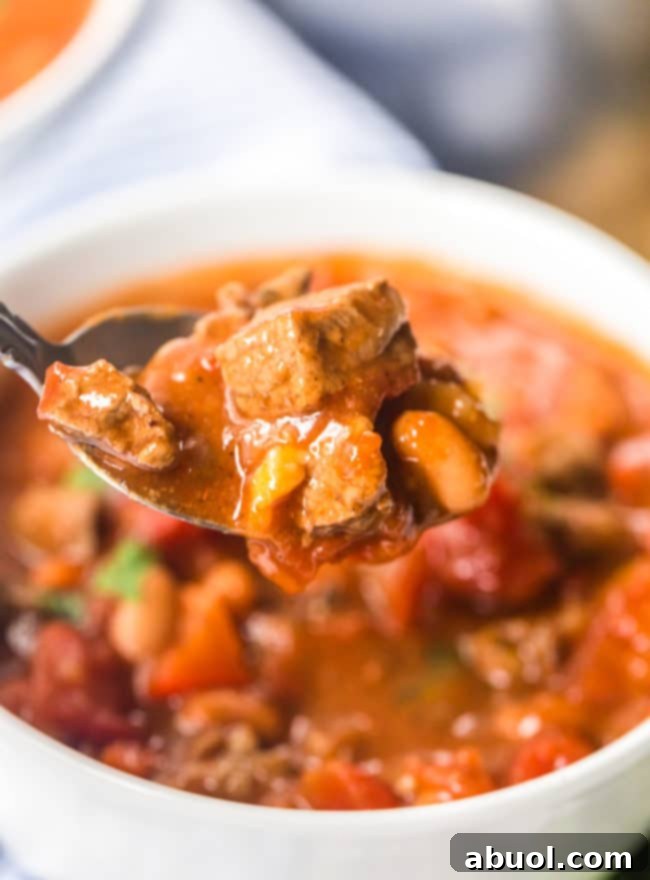 A spoon lifting a hearty mouthful of steak chili with beer, with the full bowl of chili in the background.