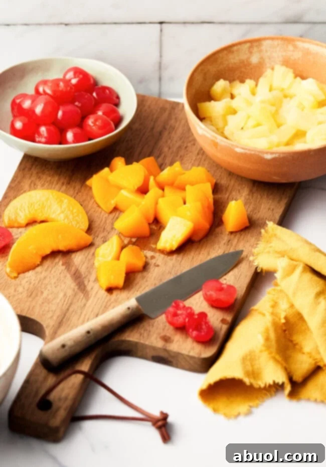 Chopped ingredients for ambrosia fruit salad on a wooden cutting board with a knife. The chopped ingredients here are peaches and maraschino cherries and pineapple. 
