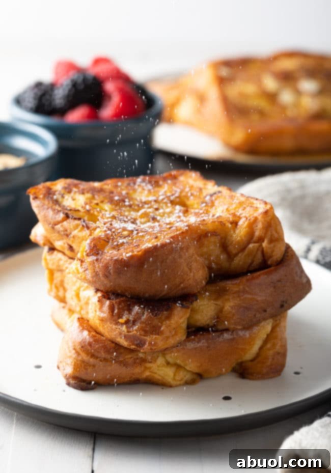 A beautifully stacked serving of golden brown French toast on a white plate, lightly dusted with powdered sugar.
