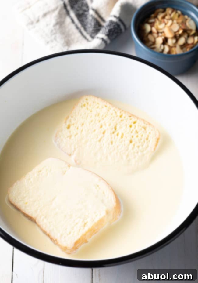 Two thick slices of brioche bread soaking deeply in a creamy custard mixture in a shallow dish.