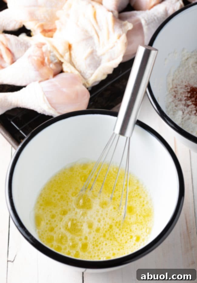 Frothy egg whites in a white mixing bowl with a metal whisk, ready for coating the chicken.