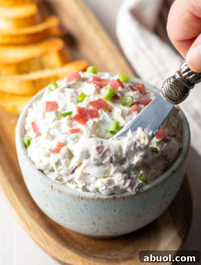 Hand holding a butter knife being dipped into the Chipped Beef Dip. A close-up showing the creamy texture.