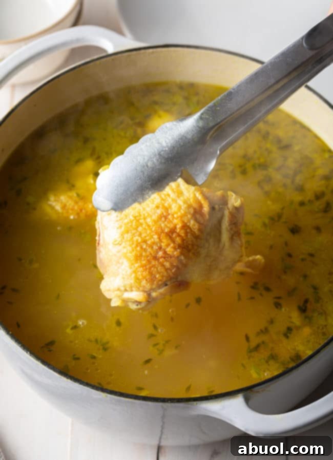 Tender chicken thighs being carefully placed into a simmering pot of chicken noodle soup.