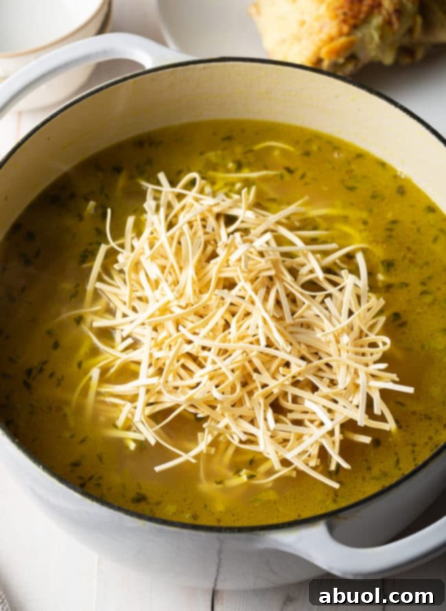 Dried Kluski egg noodles being added to a rich, simmering pot of chicken noodle soup.