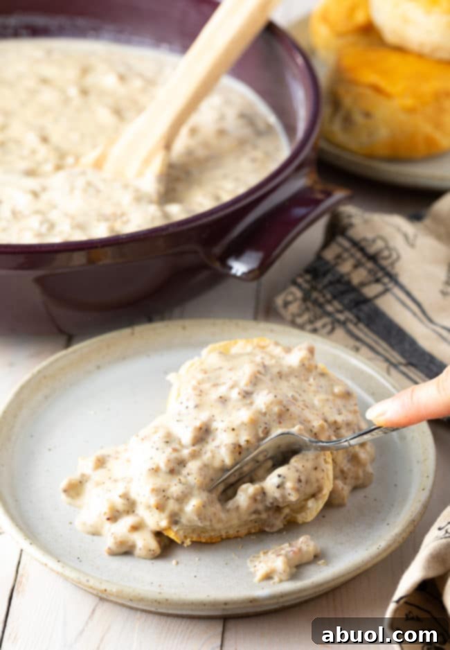Effortless Sausage Gravy 4 Hand cutting into a biscuit smothered with spicy sausage gravy on a plate.