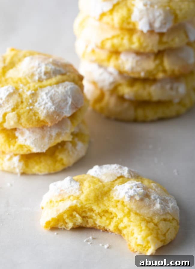 A lemon crinkle cookie with a bite taken out, revealing its soft, yellow interior. More cookies are stacked in the background, creating an appealing composition.