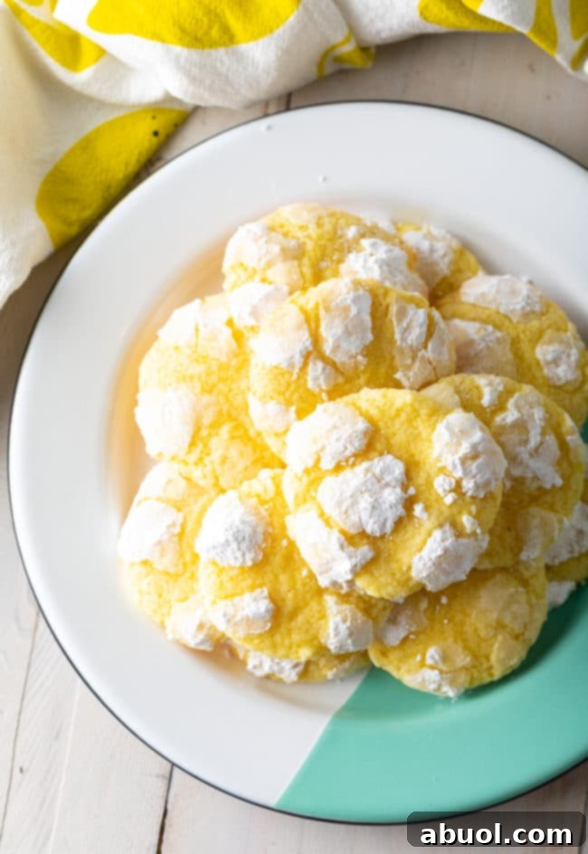 A close-up of a stack of lemon crinkle cookies, showing their perfect crackled tops and inviting yellow interior, set against a blurred background of more cookies.