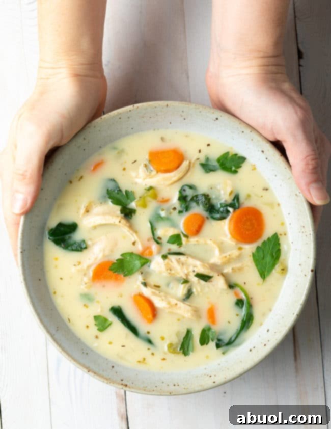 Velvety Lemon Chicken & Spinach Soup 2 Hands holding a speckled bowl filled with creamy lemon chicken soup, garnished with fresh herbs.